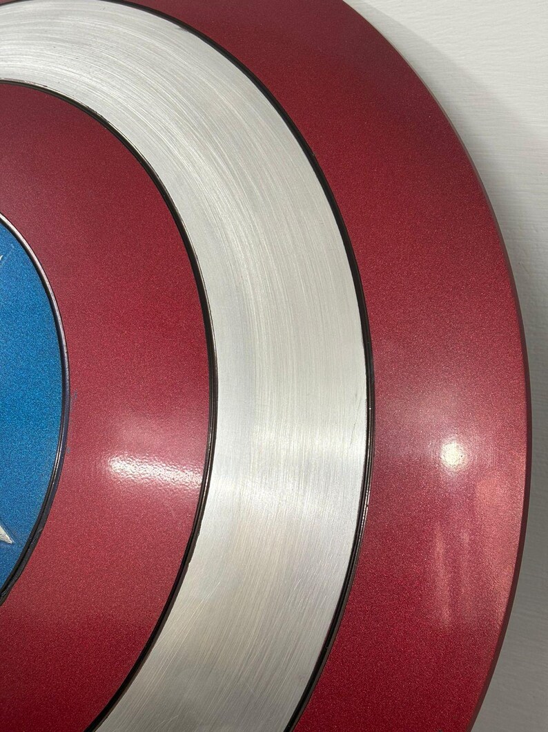 May include: A circular shield featuring concentric rings of metallic red and brushed silver, with a vibrant blue center section. The design consists of a smooth, reflective red outer ring, a textured brushed silver middle ring, and a blue inner circle.