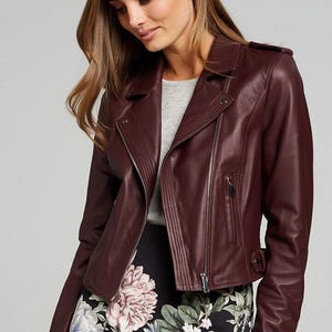 Pu&ograve; includere: Giacca in pelle bordeaux con chiusura asimmetrica con zip, revers a lancia e una piccola tasca con zip. La giacca, dalla vestibilit&agrave; aderente, &egrave; indossata sopra un top grigio e una gonna con stampa floreale.