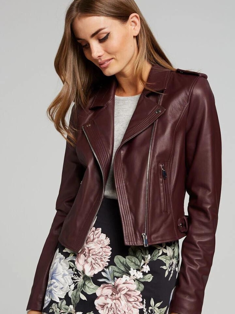 Pu&ograve; includere: Giacca in pelle bordeaux con chiusura asimmetrica con zip, revers a lancia e una piccola tasca con zip. La giacca, dalla vestibilit&agrave; aderente, &egrave; indossata sopra un top grigio e una gonna con stampa floreale.