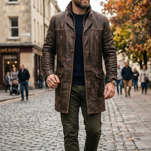 Pu&ograve; includere: Un cappotto in pelle marrone con chiusura a bottoni, due tasche sul petto e due tasche inferiori. Il cappotto &egrave; indossato sopra un maglione blu navy e pantaloni verde oliva. Un orologio &egrave; visibile al polso. La persona cammina su una strada acciottolata.