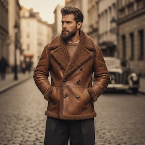 May include: A brown leather jacket with a shearling collar and lining. The double-breasted coat has large buttons and pockets. The person in the image is wearing a sweater and dark grey trousers. The background shows a cobbled street and buildings.