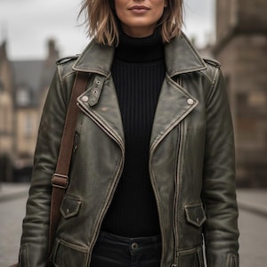 Può includere: Giacca in pelle verde oliva invecchiata in classico stile biker. La giacca presenta un colletto a revers, una cerniera asimmetrica e più tasche. La modella indossa un dolcevita nero e pantaloni neri, con una borsa a tracolla marrone.