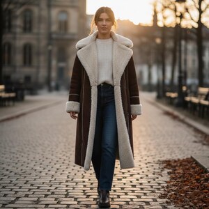 May include: A woman wearing a long, brown shearling coat with cream-colored lining and trim. The coat is open, revealing a cream-colored sweater and dark blue jeans. The woman is walking on a cobblestone street.
