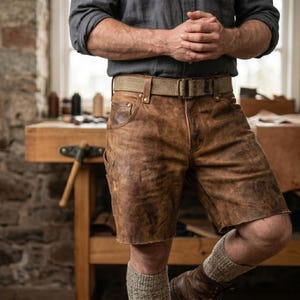 Puede incluir: Pantalones cortos de cuero marrón con acabado envejecido, con bolsillos y cinturón a juego. Los pantalones cortos se combinan con una camisa gris y calcetines, creando una estética vintage y robusta. Los pantalones cortos parecen hechos a mano.