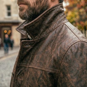 Pu&ograve; includere: Giacca di pelle marrone con finitura strutturata. La giacca ha un colletto classico e chiusura con bottoni. L'immagine mostra la spalla e la parte superiore del petto della giacca, con uno sfondo sfocato di una scena di strada.