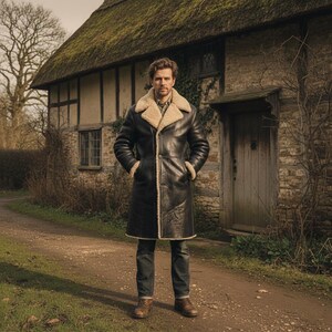 Peut inclure: Un long manteau en cuir noir avec une doublure et un col en peau de mouton cr&egrave;me. Le manteau a un design classique avec une fermeture &agrave; boutons et des poches. La personne porte un jean bleu et des bottes marron. En arri&egrave;re-plan, un cottage en pierre avec un toit de chaume.