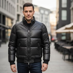 Puede incluir: Chaqueta acolchada de cuero negro con cuello alto y cierre de cremallera frontal. La chaqueta tiene un diseño acolchado y puños y dobladillo acanalados. La persona lleva vaqueros azules. El fondo es una calle de la ciudad.