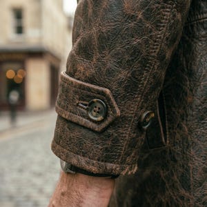 Pu&ograve; includere: Primo piano di una manica di giacca in pelle marrone. La giacca ha un aspetto strutturato e invecchiato con cuciture visibili e due bottoni marrone scuro. Il polsino presenta un dettaglio con patta abbottonata e un orologio &egrave; visibile al polso.
