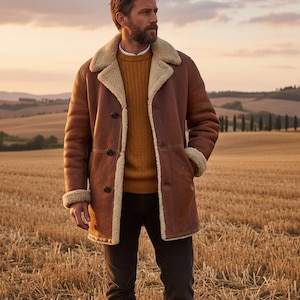 May include: A brown leather coat with a cream-colored shearling lining and collar. The coat has a button-up front and is worn over a mustard-colored sweater. The person is standing in a field of dry grass.