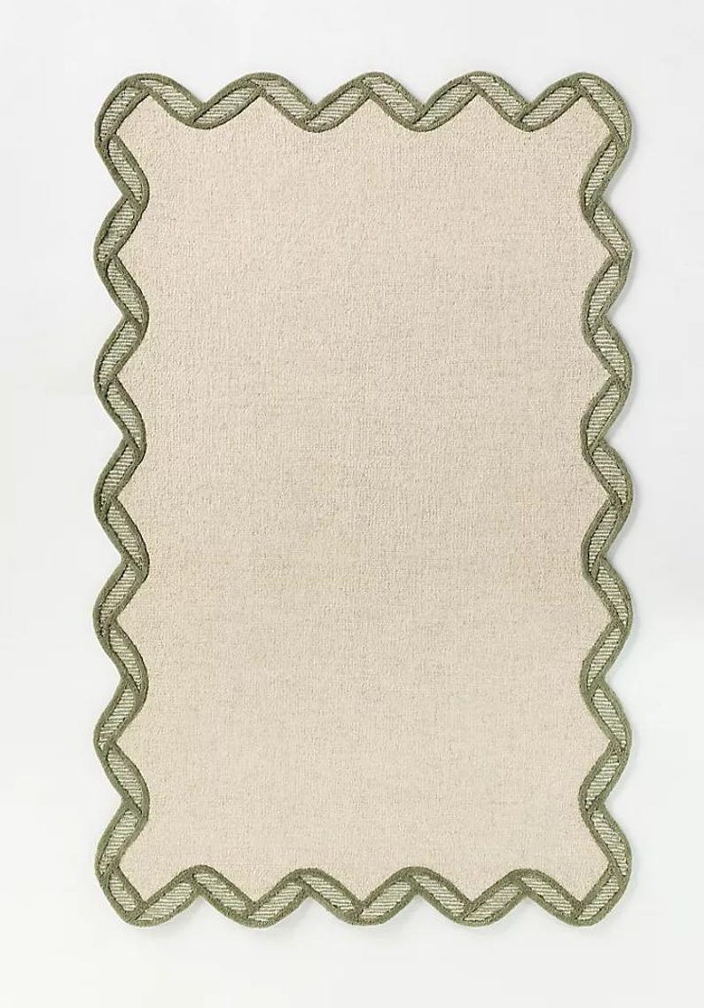 May include: A rectangular, off-white placemat with a scalloped edge. The edge is trimmed with a woven, olive green border. The placemat has a textured appearance and is set against a white background.