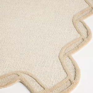 May include: A cream-colored, scalloped-edge rug with a textured surface. The rug's border is a lighter shade of beige, creating a subtle contrast. The design is simple and elegant, suitable for a bathroom or bedroom.