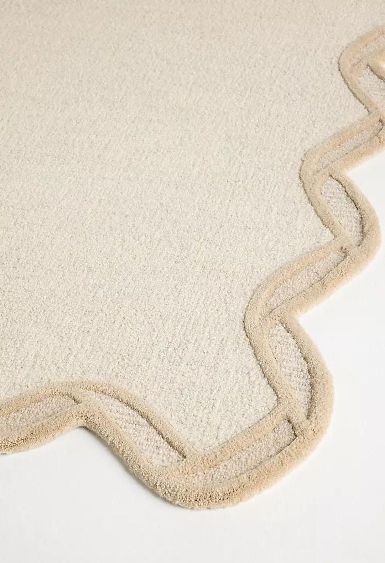 May include: A cream-colored, scalloped-edge rug with a textured surface. The rug's border is a lighter shade of beige, creating a subtle contrast. The design is simple and elegant, suitable for a bathroom or bedroom.
