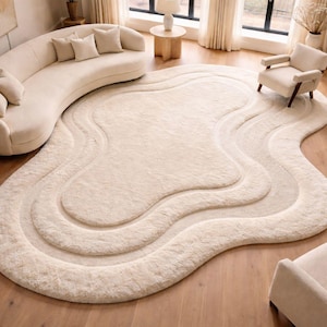 Op de afbeelding: Een crèmekleurig, abstract gevormd vloerkleed met een gelaagd ontwerp, geplaatst op een houten vloer. Het vloerkleed is omgeven door een gebogen bank, twee fauteuils en kleine bijzettafels, waardoor een gezellige woonkamer ontstaat.