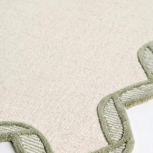 May include: Off-white rug with a scalloped edge in a light green color. The rug has a soft, textured surface. The edge features a repeating pattern of curved shapes.