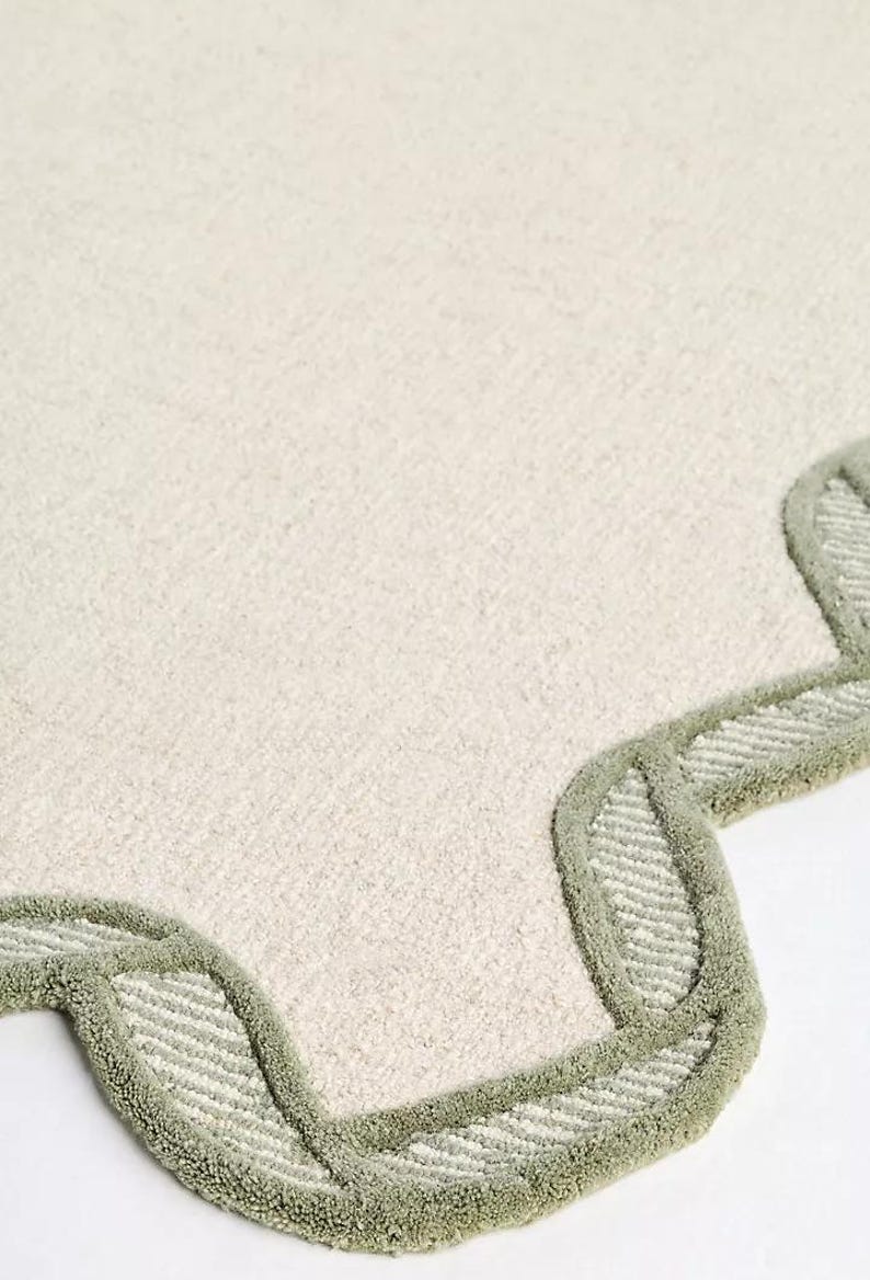 May include: Off-white rug with a scalloped edge in a light green color. The rug has a soft, textured surface. The edge features a repeating pattern of curved shapes.