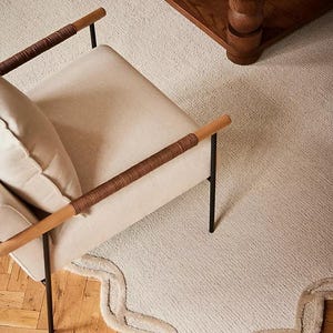 May include: A neutral-toned living room scene featuring a cream-colored chair with wooden armrests wrapped in brown woven material. A matching rug with a scalloped edge and a wooden coffee table are also visible.