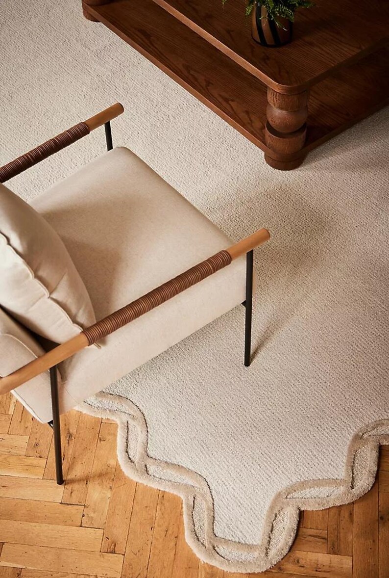 May include: A neutral-toned living room scene featuring a cream-colored chair with wooden armrests wrapped in brown woven material. A matching rug with a scalloped edge and a wooden coffee table are also visible.