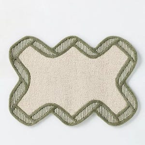 May include: A beige bath mat with a unique, wavy shape. The mat has a light green border with a striped pattern. The center of the mat is a solid beige color, creating a simple yet stylish design.
