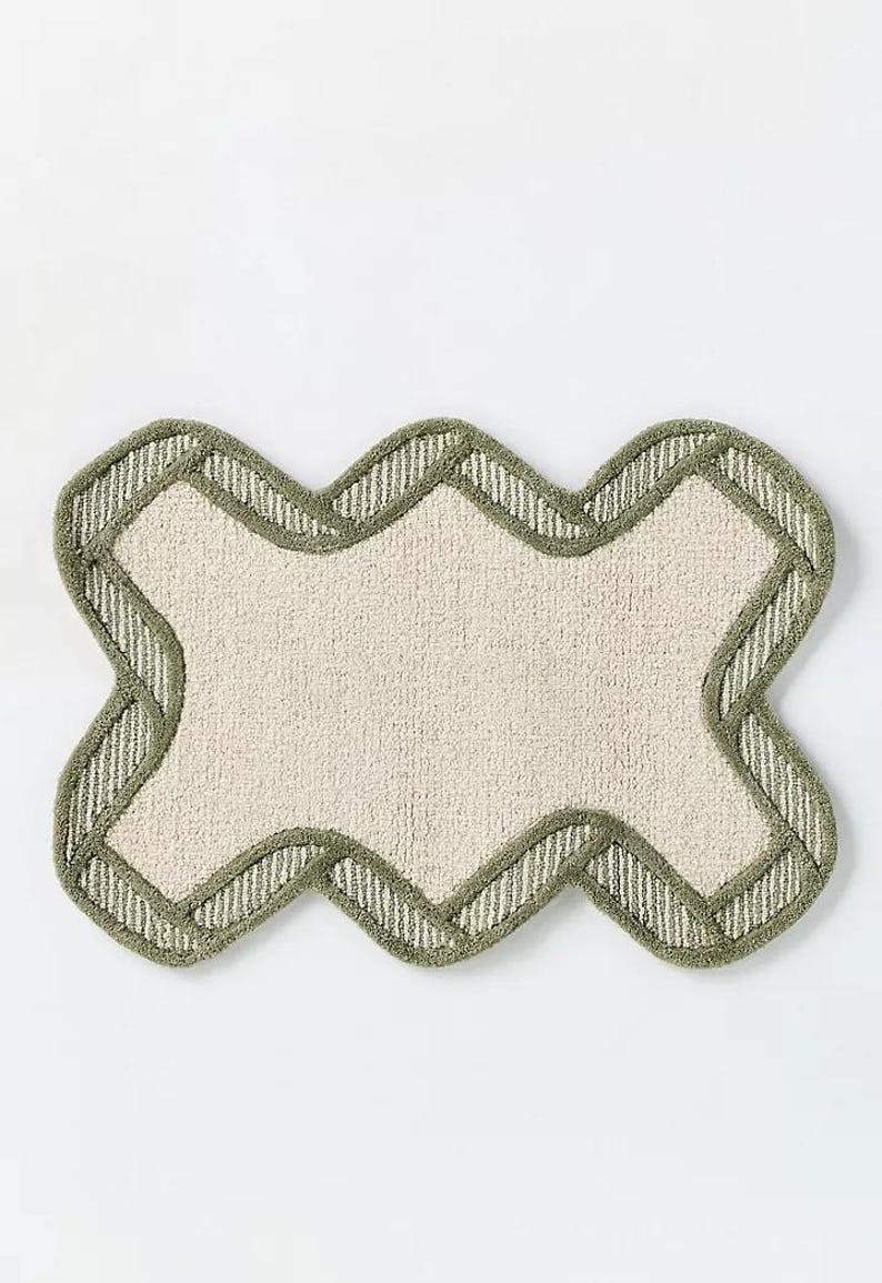May include: A beige bath mat with a unique, wavy shape. The mat has a light green border with a striped pattern. The center of the mat is a solid beige color, creating a simple yet stylish design.