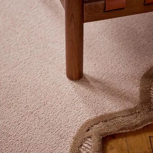 May include: A close-up of a light pink carpet with a brown, wavy border. A wooden chair with leather straps and a black cushion is partially visible. The carpet has a textured surface.