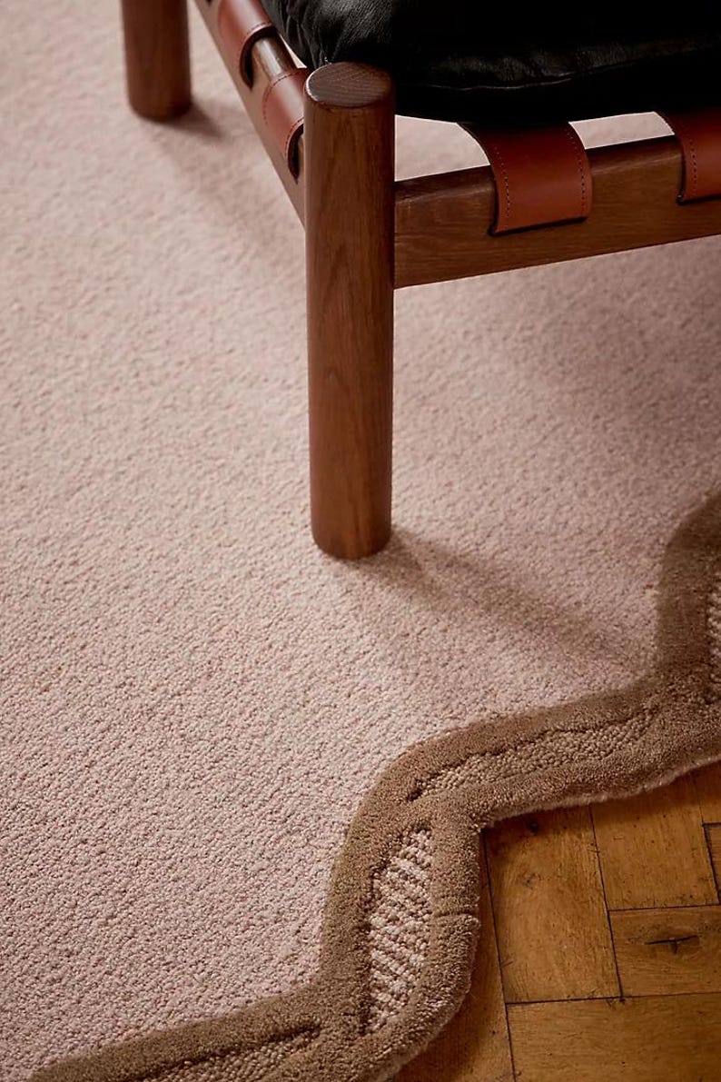 May include: A close-up of a light pink carpet with a brown, wavy border. A wooden chair with leather straps and a black cushion is partially visible. The carpet has a textured surface.