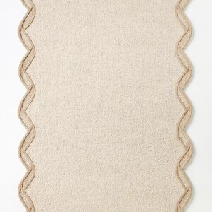 May include: A rectangular beige rug with a scalloped edge. The rug has a textured surface and a light brown border that follows the wavy outline. The rug is a neutral color and would fit in many home decor styles.