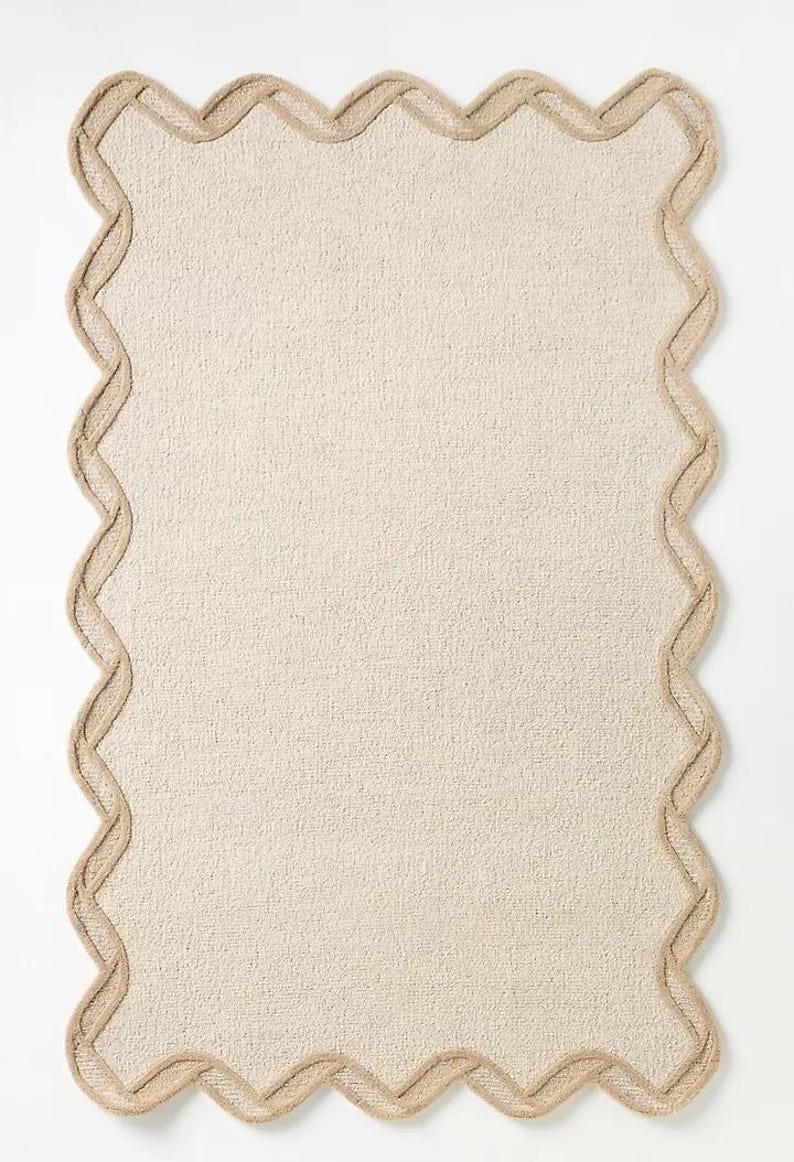 May include: A rectangular beige rug with a scalloped edge. The rug has a textured surface and a light brown border that follows the wavy outline. The rug is a neutral color and would fit in many home decor styles.