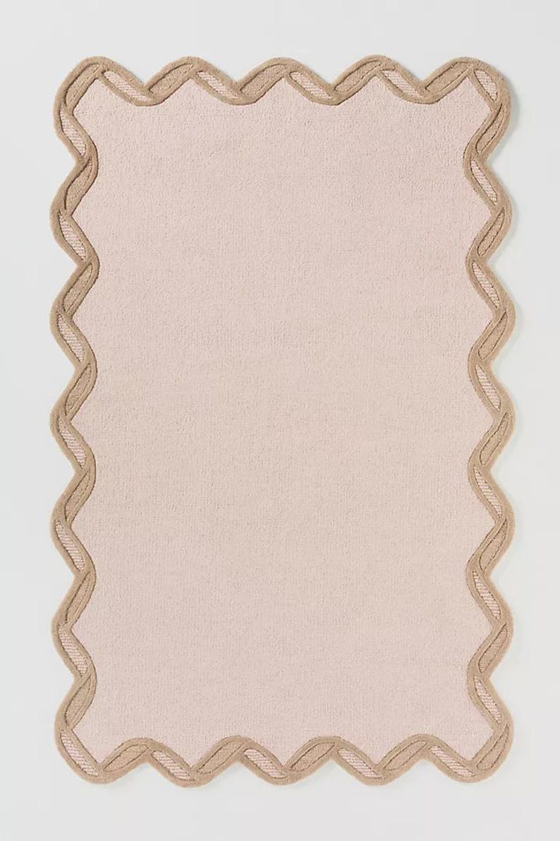 May include: A rectangular, blush-colored rug with a scalloped edge. The border is a light tan color, creating a contrasting frame. The rug has a soft, textured appearance, suitable for home decor.