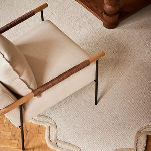 May include: An overhead shot of a cream-colored rug with a scalloped edge, a beige armchair with brown woven armrests, and a dark wood coffee table. The rug is on a light wood floor.