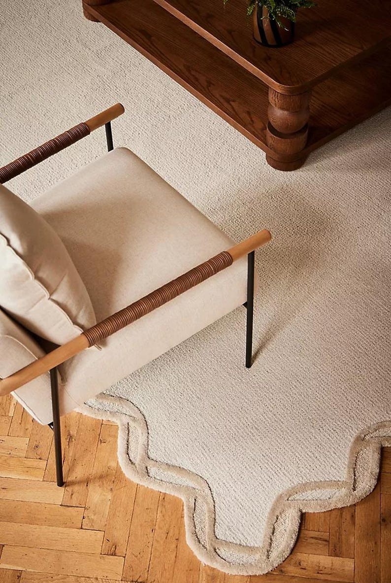 May include: An overhead shot of a cream-colored rug with a scalloped edge, a beige armchair with brown woven armrests, and a dark wood coffee table. The rug is on a light wood floor.