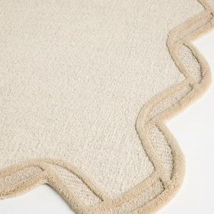 May include: Off-white, textured rug with a scalloped edge in a light beige color. The rug has a soft, plush appearance, and the border adds a decorative element. The rug is suitable for a bathroom or bedroom.