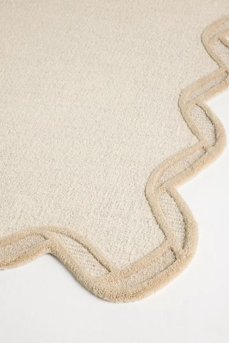 May include: Off-white, textured rug with a scalloped edge in a light beige color. The rug has a soft, plush appearance, and the border adds a decorative element. The rug is suitable for a bathroom or bedroom.