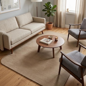 May include: A living room set featuring a beige textured sofa, two gray upholstered armchairs with wooden frames, and a round wooden coffee table. The table holds a book, a small bowl, and a candle. A large beige area rug sits on the floor.