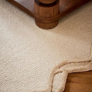 May include: Close-up of a wooden table leg with a rounded design, set on a cream-colored carpet. The carpet has a textured surface and a decorative border in a lighter shade of beige. The table leg is made of dark wood.