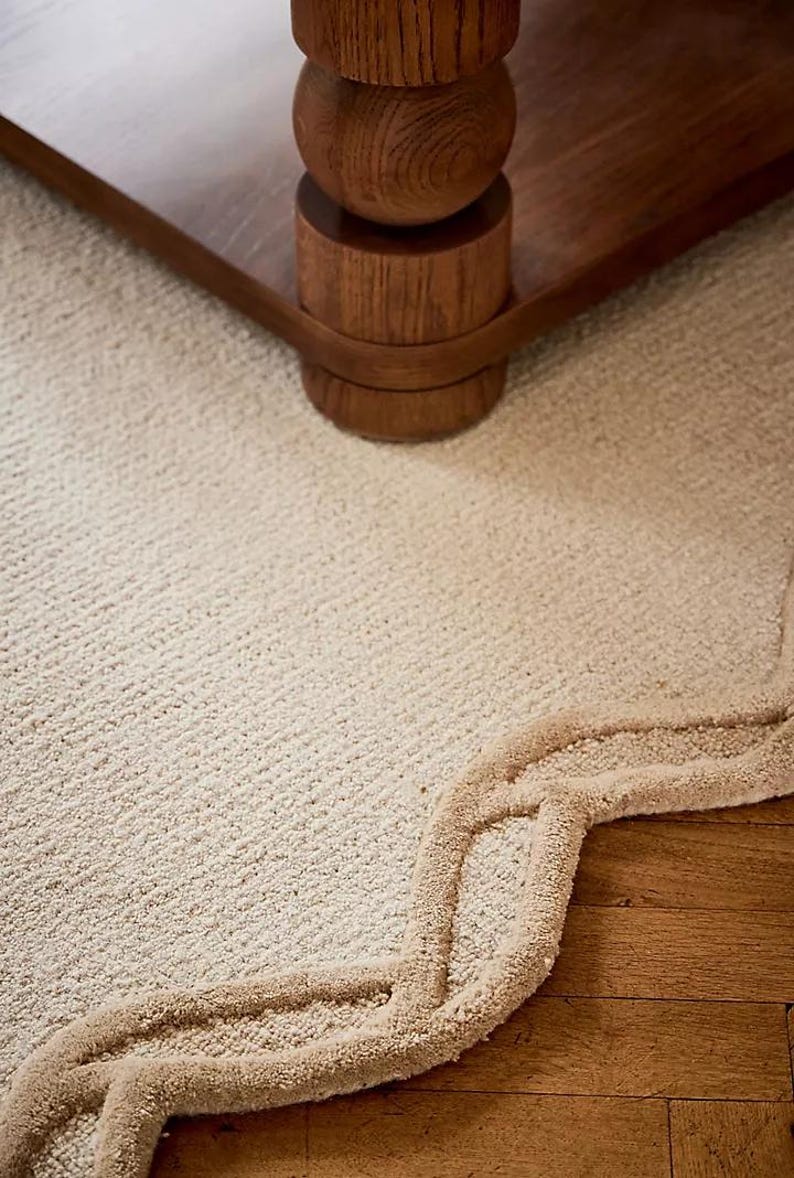 May include: Close-up of a wooden table leg with a rounded design, set on a cream-colored carpet. The carpet has a textured surface and a decorative border in a lighter shade of beige. The table leg is made of dark wood.