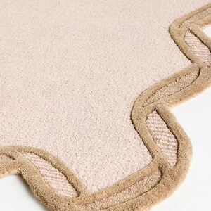 May include: A light pink rug with a scalloped edge. The border is a darker tan color, creating a contrasting outline. The rug has a soft, textured appearance, suitable for a bedroom or living room.