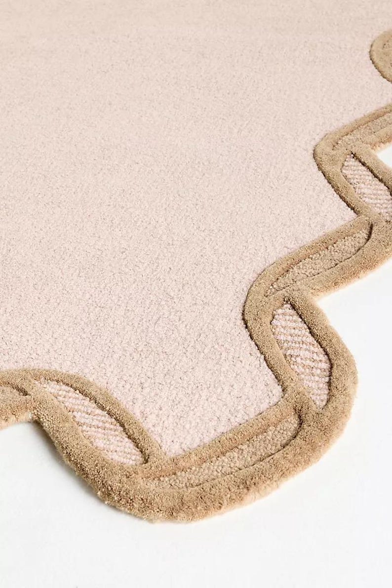 May include: A light pink rug with a scalloped edge. The border is a darker tan color, creating a contrasting outline. The rug has a soft, textured appearance, suitable for a bedroom or living room.