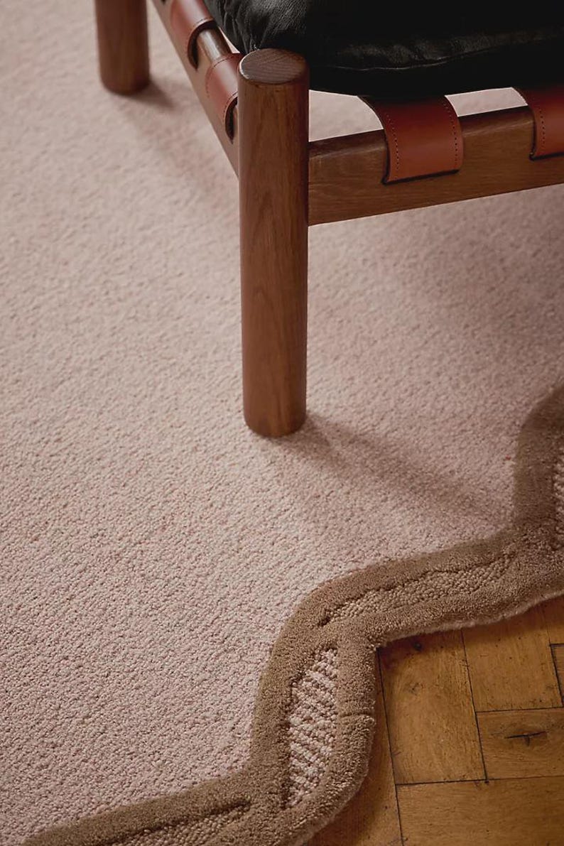 May include: A close-up of a light pink rug with a wavy, brown border. A wooden chair with leather straps and a black cushion is partially visible. The rug has a textured surface.