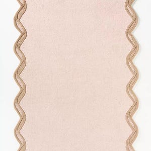 May include: A rectangular, light pink placemat with a scalloped edge. The edge is a contrasting tan color with a woven design. The placemat is made of a textured fabric and is set against a white background.