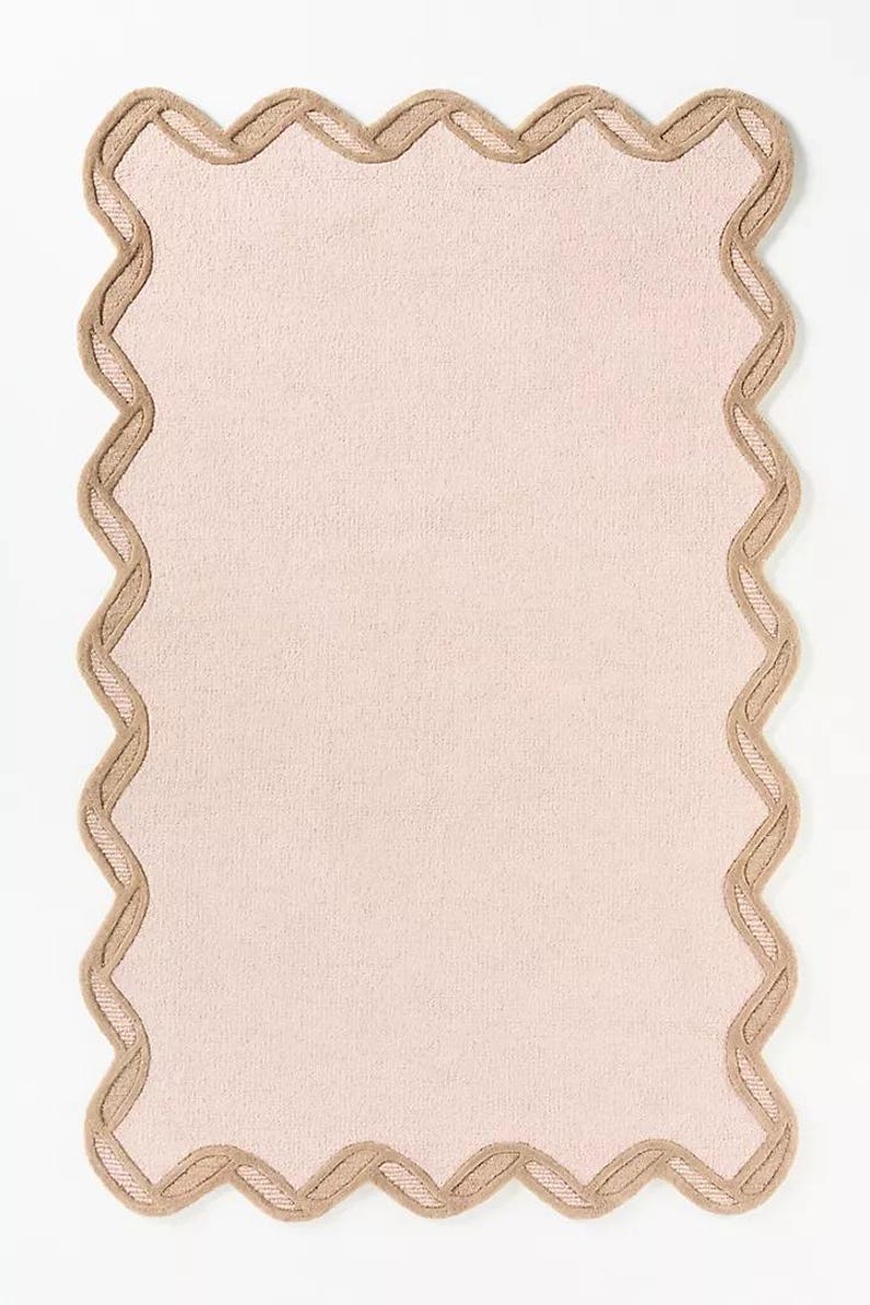 May include: A rectangular, light pink placemat with a scalloped edge. The edge is a contrasting tan color with a woven design. The placemat is made of a textured fabric and is set against a white background.