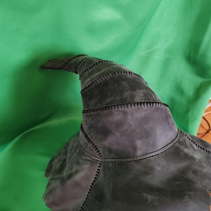 Op de afbeelding: Een donkergrijze leren heksenhoed met een puntige kroon en een brede, gekartelde rand. De hoed is gemaakt van verschillende stukken leer die met zwart garen aan elkaar zijn gestikt. De achtergrond is een heldergroene stof.