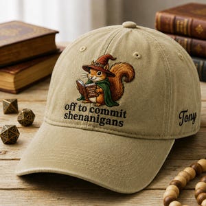 May include: Tan baseball cap featuring a cartoon squirrel in a wizard hat and cape, reading a book. The text "off to commit shenanigans" is below the squirrel. The name "Tony" is embroidered on the side. A fun, whimsical accessory.