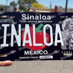 May include: Black license plate featuring the word "SINALOA" in large white letters, with a red tomato graphic. The plate also includes the text "MÉXICO" and an image of a religious figure. The plate is held by a hand.