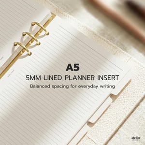 Può includere: Un inserto per planner A5 aperto con pagine a righe da 5 mm. Il testo sulla pagina recita "5MM LINED PLANNER INSERT" e "Balanced spacing for everyday writing". L'agenda ha anelli color oro ed è appoggiata su una superficie dai toni neutri.