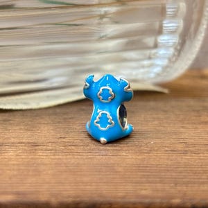 May include: A blue charm with a silver outline and a cartoon-like design. The charm has a light blue body with silver accents and a dark blue center. The charm is on a wooden surface.