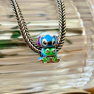 May include: A colourful charm featuring a cartoon character in blue, green, and purple enamel. The charm is attached to a silver chain. The charm is a small, detailed piece of jewellery.