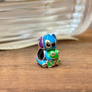 May include: A detailed metal charm depicting a blue alien character with purple ears and a green frog. The charm is brightly coloured. This charm is a collectible item.