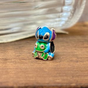 May include: A small, colourful charm featuring the character Stitch from the animated film. The charm is primarily blue with purple ears, a green belly, and black eyes. The charm is sitting on a wooden surface.