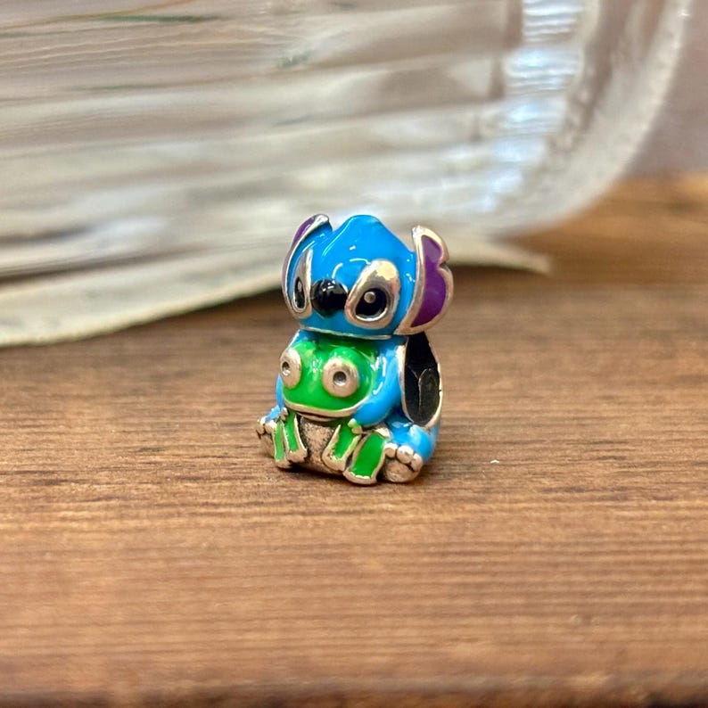 May include: A small, colourful charm featuring the character Stitch from the animated film. The charm is primarily blue with purple ears, a green belly, and black eyes. The charm is sitting on a wooden surface.