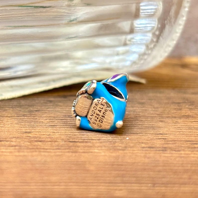 May include: A blue and silver charm with the text "PANDORA S925 ALE &copy; Disney". The charm is in the shape of a character with a blue body, silver accents, and purple details. The charm is on a wooden surface.
